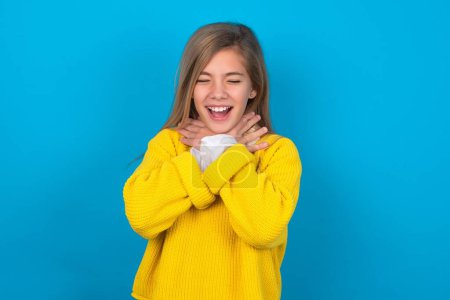 Foto de Chica adolescente caucásica usando suéter amarillo sobre la pared azul gritando sofocar porque estrangulamiento doloroso. Problema de salud. Concepto de asfixia y suicidio. - Imagen libre de derechos