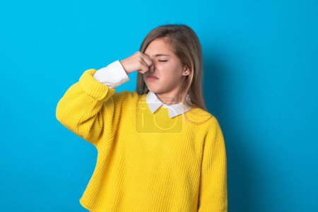 Téléchargez les photos : Adolescente caucasienne portant un pull jaune sur un mur bleu sentant quelque chose de puant et dégoûtant, odeur intolérable, retenant la respiration avec les doigts sur le nez. Mauvaise odeur - en image libre de droit