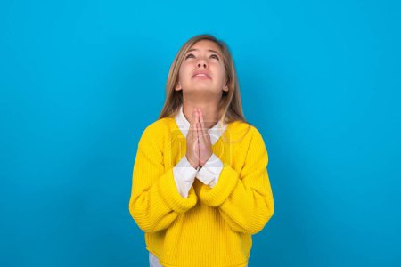 Téléchargez les photos : Adolescent caucasien fille portant un pull jaune sur le mur bleu mendier et prier avec les mains ainsi que l'expression d'espoir sur le visage très émotionnel et inquiet. Mon Dieu ! - en image libre de droit