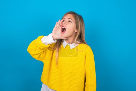 Foto de Chica adolescente caucásica usando suéter amarillo sobre la pared azul gritando y gritando fuerte a un lado con la mano en la boca. Concepto de comunicación. - Imagen libre de derechos
