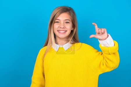 Téléchargez les photos : Adolescent caucasien fille portant un pull jaune sur le mur bleu geste souriant et confiant avec la main faisant signe de petite taille avec les doigts à la recherche et la caméra. Concept de mesure - en image libre de droit