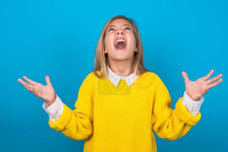 Photo for Caucasian teen girl wearing yellow sweater over blue wall crazy and mad shouting and yelling with aggressive expression and arms raised. Frustration concept. - Royalty Free Image