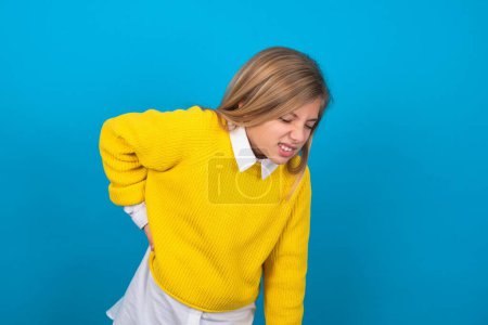 Foto de Chica adolescente caucásica usando suéter amarillo sobre la pared azul Sufrimiento de dolor de espalda, tocando la espalda con la mano, dolor muscular - Imagen libre de derechos