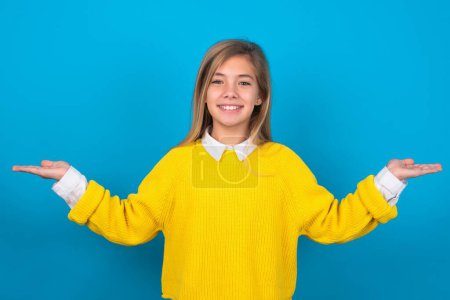 Téléchargez les photos : Gai gai optimiste caucasien adolescent fille portant jaune pull sur bleu mur tenant deux paumes copie espace - en image libre de droit