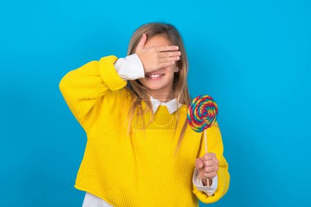 Photo for Caucasian teen girl wearing yellow sweater over blue wall smiling and laughing with hand on face covering eyes for surprise. Blind concept. - Royalty Free Image