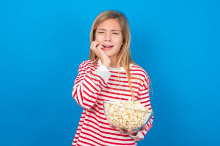Photo for Doleful desperate crying blonde teen girl wearing striped t-shirt over blue wall looks stressfully, frowns face, feels lonely and anxious - Royalty Free Image