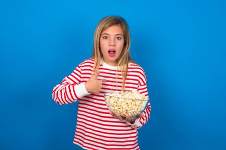 Photo for Embarrassed blonde teen girl wearing striped t-shirt over blue wall indicates at herself with puzzled expression, being shocked to be chosen to participate in competition, hesitates about something - Royalty Free Image