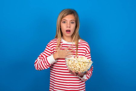 Photo for Scared blonde teen girl wearing striped t-shirt over blue wall looks with frightened expression, keeps hands on chest, being puzzled to notice something strange, People, hush reaction and emotions. - Royalty Free Image