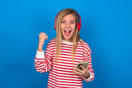 Foto de Chica adolescente rubia positiva con camiseta a rayas sobre la pared azul sostiene el teléfono celular moderno conectado a los auriculares, aprieta el puño de las buenas emociones, exclama con alegría, - Imagen libre de derechos