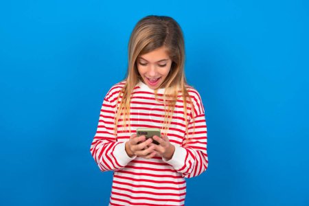 Foto de Rubia adolescente chica usando rayas camiseta sobre azul pared usando teléfono móvil chat tiempo libre . - Imagen libre de derechos
