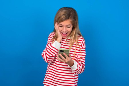 Photo for Blonde teen girl wearing striped t-shirt over blue wall holding in hands cell reading sms using new app 5g - Royalty Free Image