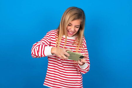Téléchargez les photos : Blonde adolescent fille portant rayé t-shirt sur le mur bleu tenant dans les mains cellulaire jouer à des jeux vidéo ou bavarder - en image libre de droit