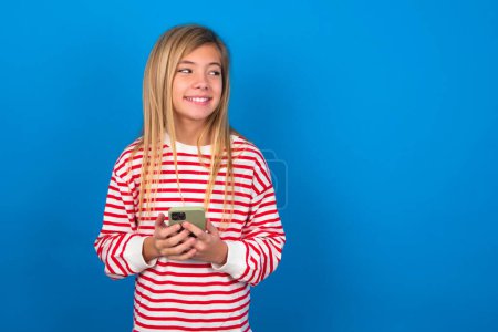 Foto de Rubia adolescente chica usando rayas camiseta sobre azul pared sostener teléfono manos leer buena juventud noticias mirar vacío espacio anuncio - Imagen libre de derechos