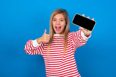 Photo for Portrait blonde teen girl wearing striped t-shirt over blue wall holding in hands cell showing giving black screen thumb up - Royalty Free Image