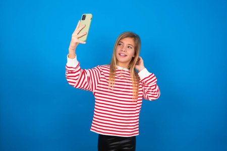 Foto de Rubia adolescente chica usando rayas camiseta sobre la pared azul sonriendo y tomando una selfie listo para publicarlo en sus redes sociales. - Imagen libre de derechos