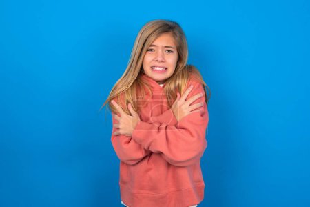 Foto de Desesperado hermosa chica adolescente caucásica con suéter rosa sobre la pared azul tiembla y se siente frío, se abraza a sí mismo para calentar o se siente asustado nota algo aterrador. - Imagen libre de derechos