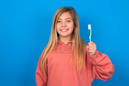 Foto de Hermosa chica adolescente caucásica con suéter rosa sobre la pared azul sosteniendo un cepillo de dientes y sonriendo. Concepto de salud dental. - Imagen libre de derechos