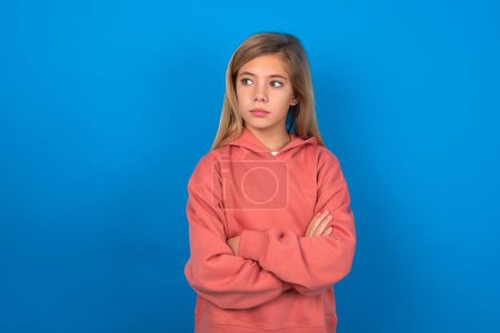 Photo for Charming thoughtful beautiful caucasian teen girl wearing pink sweater over blue wall stands with arms folded concentrated somewhere with pensive expression thinks what to do - Royalty Free Image