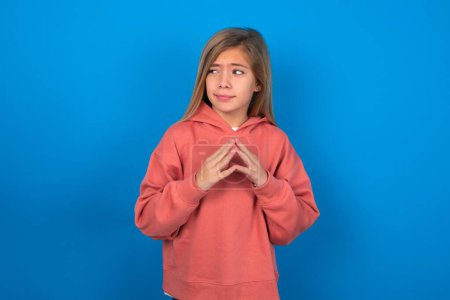 Photo for Beautiful caucasian teen girl wearing pink sweater over blue wall steepled fingers and looks mysterious aside has great evil plan in mind - Royalty Free Image