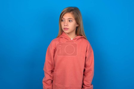 Photo for Beautiful caucasian teen girl wearing pink sweater over blue wall stares aside with wondered expression has speechless expression. Embarrassed model looks in surprise - Royalty Free Image