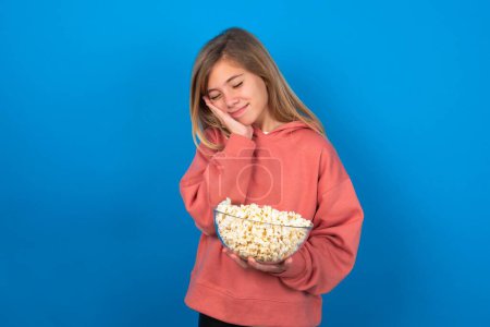 Photo for Beautiful caucasian teen girl wearing pink sweater over blue wall leans on pressed palms closes eyes and has pleasant smile dreams about something - Royalty Free Image
