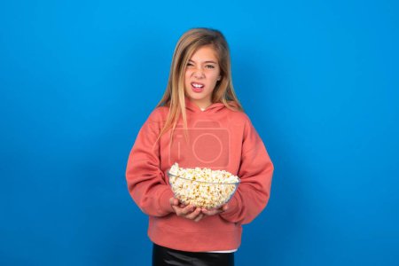 Photo for Portrait of dissatisfied beautiful caucasian teen girl wearing pink sweater over blue wall smirks face, purses lips and looks with annoyance at camera, discontent hearing something unpleasant - Royalty Free Image