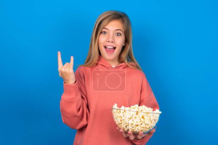 Photo for Beautiful caucasian teen girl wearing pink sweater over blue wall makes rock n roll sign looks self confident and cheerful enjoys cool music at party. Body language concept. - Royalty Free Image
