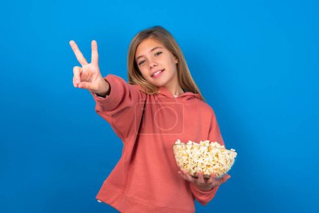 Téléchargez les photos : Belle adolescente caucasienne portant un pull rose sur un mur bleu souriant et regardant heureux, insouciant et positif, geste de victoire ou de paix avec une main - en image libre de droit