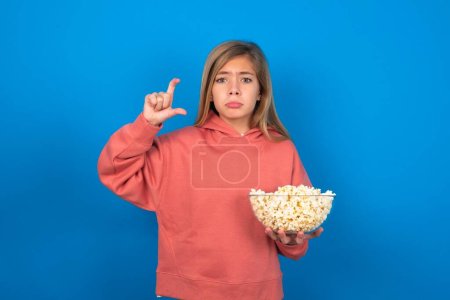 Photo for Beautiful caucasian teen girl wearing pink sweater over blue wall purses lip and gestures with hand, shows something very little. - Royalty Free Image
