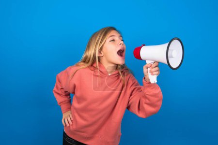 Photo for Funny beautiful caucasian teen girl wearing pink sweater over blue wall. People sincere emotions lifestyle concept. Mock up copy space. Screaming in megaphone. - Royalty Free Image