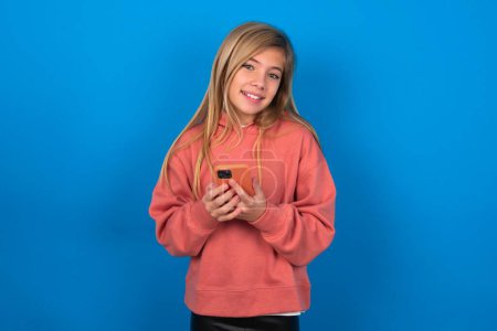 Foto de Hermosa chica adolescente caucásica con suéter rosa sobre la pared azul simulan espacio de copia. Usando el teléfono móvil, escribiendo el mensaje sms - Imagen libre de derechos