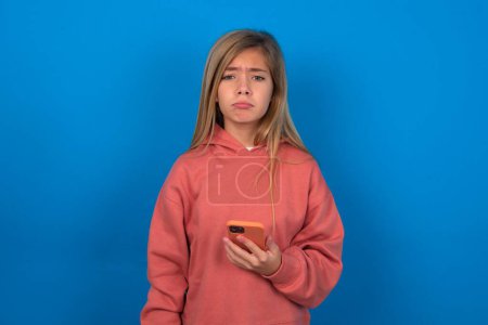 Foto de Molesto insatisfecho hermosa chica adolescente caucásica con suéter rosa sobre la pared azul utiliza la aplicación de software móvil y la información de navegación en Internet, sostiene la mano móvil moderna - Imagen libre de derechos
