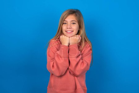 Téléchargez les photos : Satisfait belle adolescente caucasienne portant un pull rose sur un mur bleu touche menton avec les deux mains, sourit agréablement, se réjouit bonne journée avec l'amant - en image libre de droit