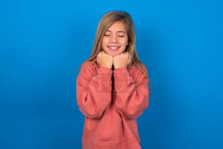 Téléchargez les photos : Joyeux beau caucasien adolescent fille portant un pull rose sur le mur bleu a timide expression satisfaite, sourit largement, montre les dents blanches, les émotions des gens - en image libre de droit