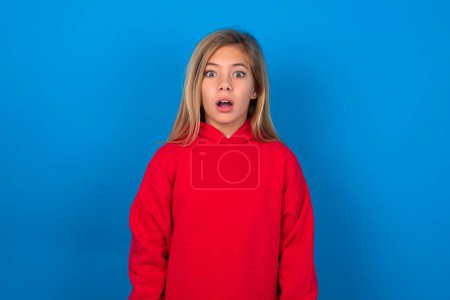 Foto de Sorprendido chica adolescente rubia con suéter rojo sobre la pared azul mira fijamente ojos con micrófonos mantiene la boca abierta ha sorprendido a la expresión. Concepto de Omg - Imagen libre de derechos