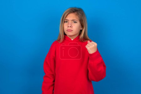 Foto de Rubia adolescente chica usando rojo suéter sobre azul pared muestra puño ha molesto expresión de la cara va a la venganza o amenazar a alguien hace mirada seria. Te mostraré quién es el jefe. - Imagen libre de derechos