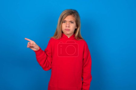 Foto de Chica adolescente rubia seria usando suéter rojo sobre la pared azul sonríe puntos de la cara en el espacio de copia muestra algo desagradable. Mira este anuncio. Gran concepto de precio - Imagen libre de derechos