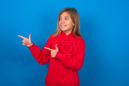Photo for Optimistic blonde teen girl wearing red sweater over blue wall points with both hands and  looking at empty space. - Royalty Free Image