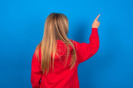 Photo for Blonde teen girl wearing red sweater over blue wall pointing to object on copy space, rear view. Turn your back - Royalty Free Image