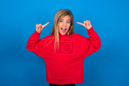 Téléchargez les photos : Joyeux adolescent blonde fille portant un pull rouge sur le mur bleu démontrant la coiffure - en image libre de droit