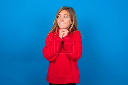 Téléchargez les photos : Curieux adolescent blonde fille portant un pull rouge sur le mur bleu garde les mains sous les lèvres de morsures de menton et regarde avec intérêt de côté. - en image libre de droit