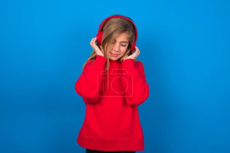 Téléchargez les photos : Adolescente blonde agréable portant un pull rouge sur le mur bleu aime écouter mélodie agréable garde les mains sur casque stéréo ferme les yeux. Passer du temps libre avec la musique - en image libre de droit