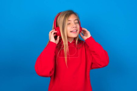 Téléchargez les photos : Blonde adolescent fille portant un pull rouge sur le mur bleu avec des écouteurs sur la tête, écoute de la musique, profiter de la chanson préférée avec les yeux fermés, tenant la main sur casque. - en image libre de droit
