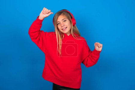 Foto de Chica adolescente rubia despreocupada usando suéter rojo sobre la pared azul con sonrisa dentada levanta brazos baila movimientos despreocupados con el ritmo de la música escucha música de la lista de reproducción a través de auriculares - Imagen libre de derechos