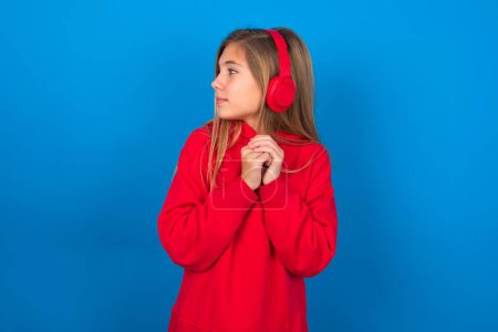 Téléchargez les photos : Blonde adolescent fille portant pull rouge sur le mur bleu porte casque stéréo écouter de la musique concentrée et regarder de côté avec intérêt. - en image libre de droit