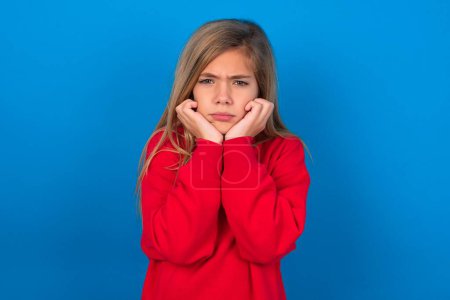 Foto de Retrato de chica adolescente rubia triste usando suéter rojo sobre la cara de manos de pared azul - Imagen libre de derechos