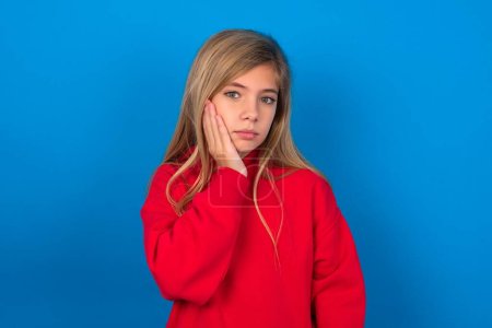 Téléchargez les photos : Photo de jeune fille blonde portant un pull rouge sur le mur bleu profiter d'une peau fraîche parfaite et lisse toucher le visage - en image libre de droit