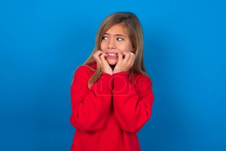 Photo for Terrified blonde teen girl wearing red sweater over blue wall looks empty space home alone moonless night - Royalty Free Image