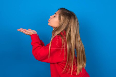 Foto de Perfil vista lateral, retrato de atractiva chica adolescente rubia usando suéter rojo sobre pared azul enviando beso de aire - Imagen libre de derechos