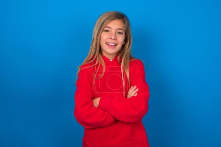 Téléchargez les photos : Portrait de charmante jeune fille blonde portant un pull rouge sur le mur bleu debout avec confiance souriant avec les mains pliées - en image libre de droit